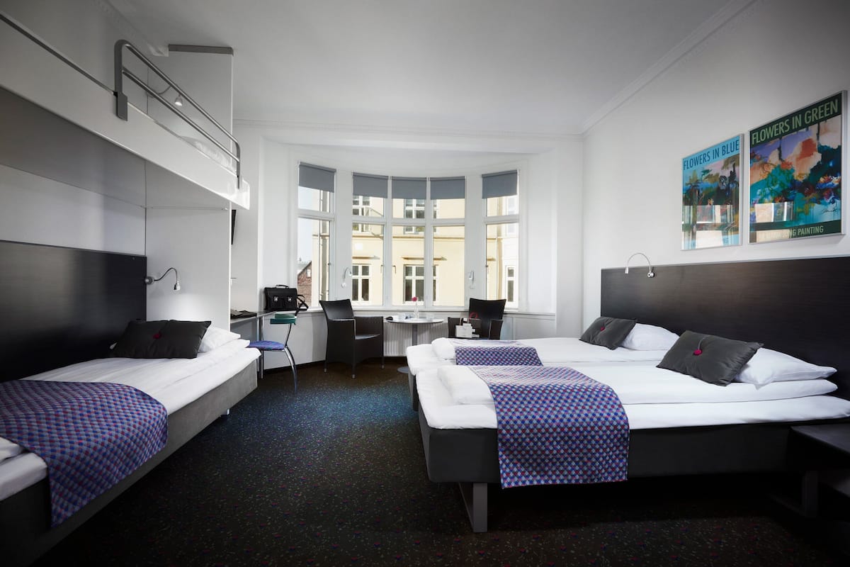 Habitación de hotel con dos camas, un escritorio y una silla. Amplias ventanas ofrecen vistas de edificios cercanos.