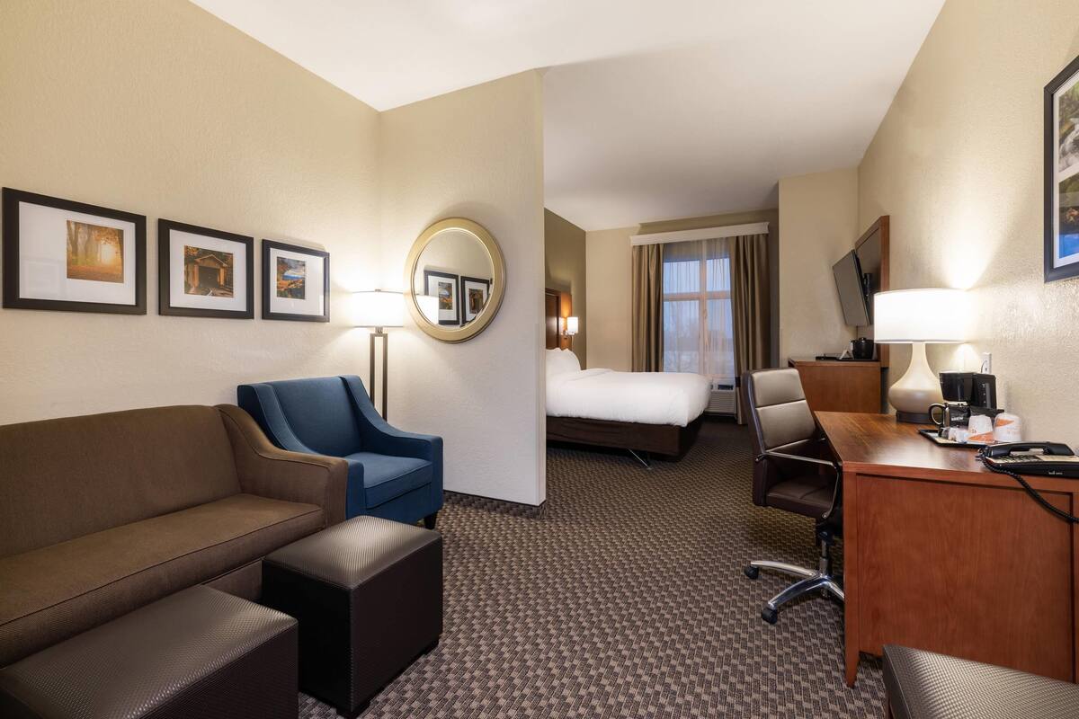 A hotel room with a bed, a desk with a lamp, a blue armchair, a brown sofa, and a small table.