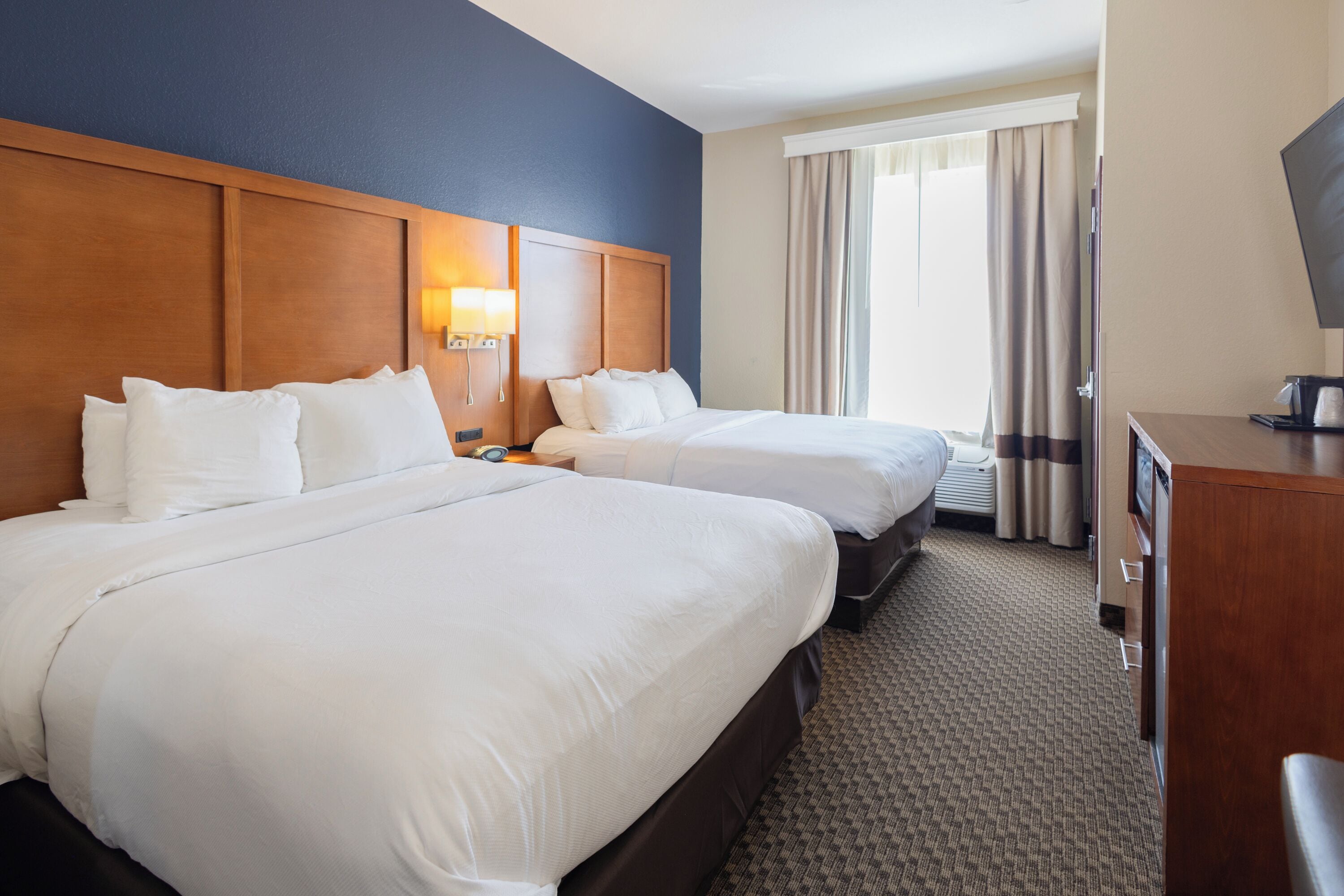 A hotel room with two beds, a wooden headboard, a nightstand with a lamp, a desk with a computer, and a window with curtains.