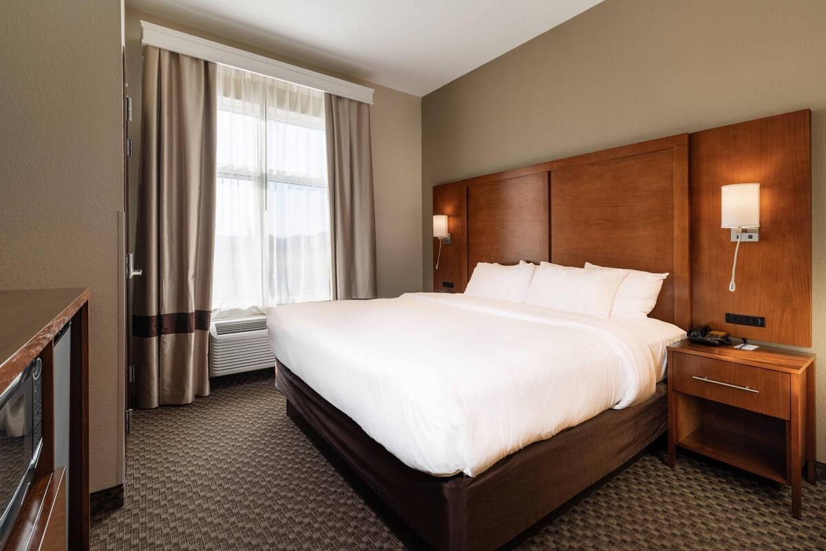 A hotel room with a large bed, a bedside table with a lamp, a desk with a telephone, and a window with curtains.