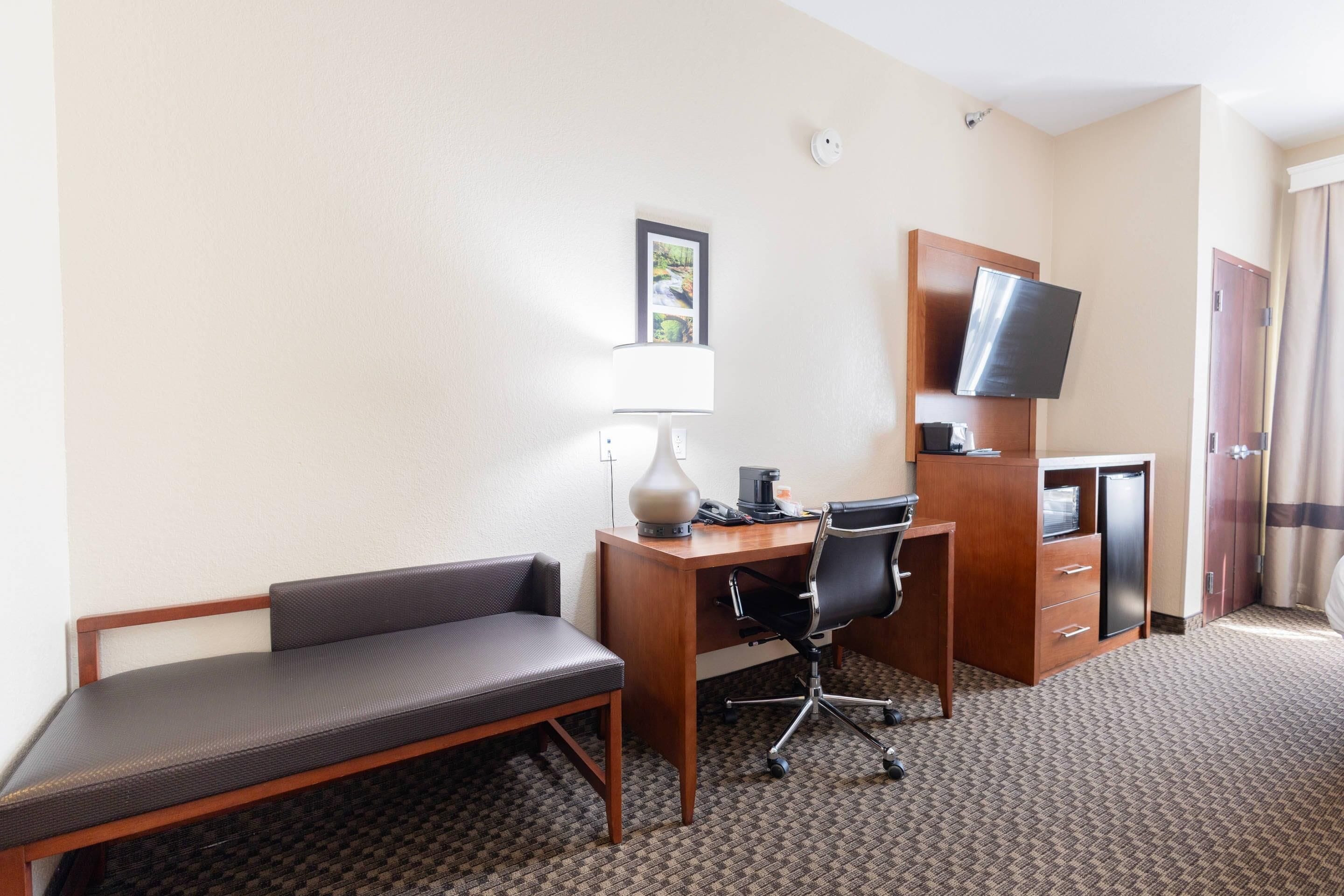 A hotel room with a desk, chair, lamp, television, and a minibar.