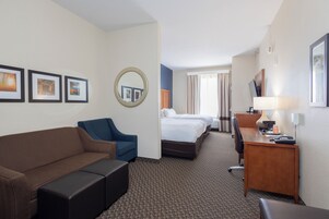 A hotel room with a brown sofa, a blue armchair, a desk with a lamp, a bed, and a large mirror.
