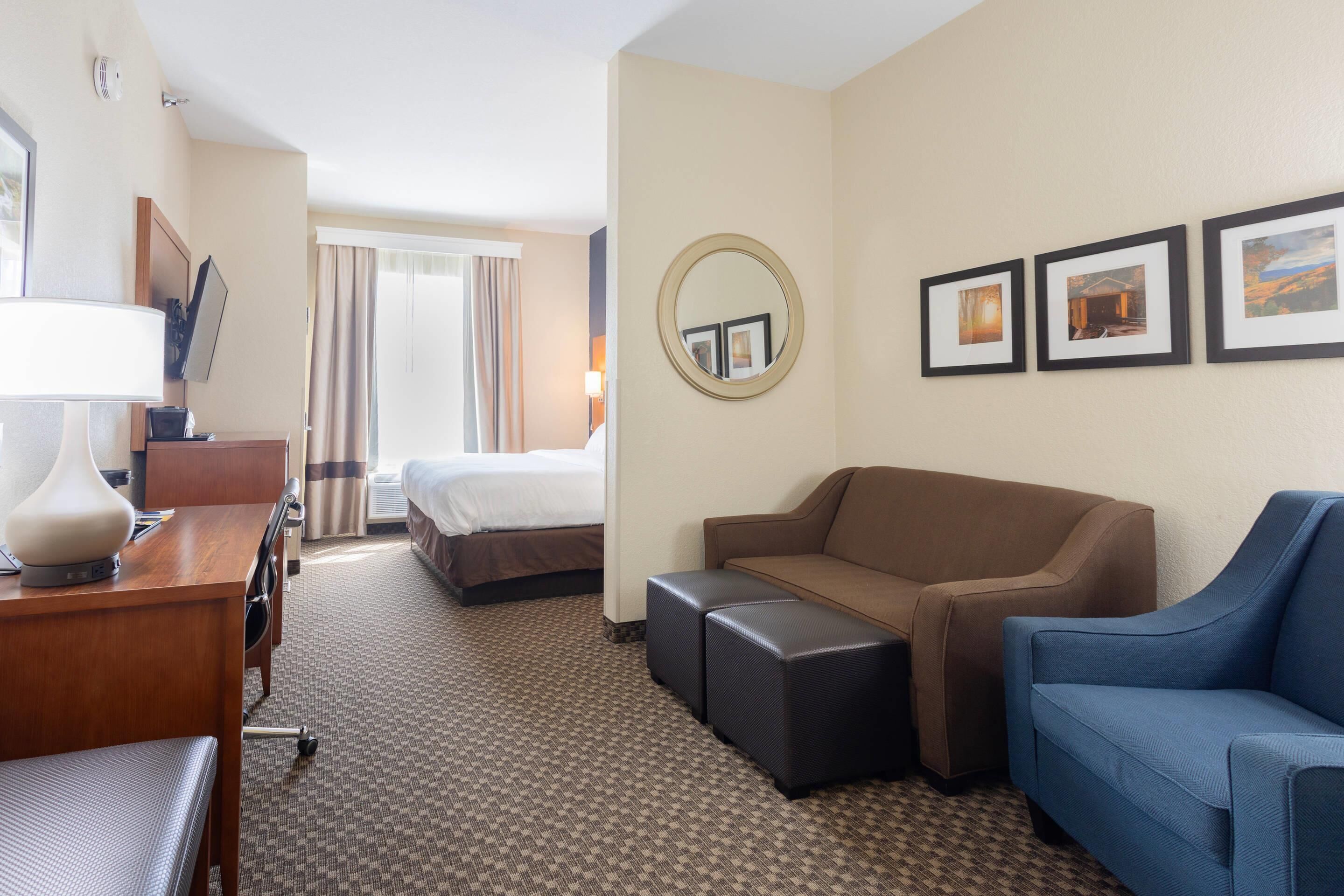 A hotel room with a bed, a desk with a lamp, a sofa, and a blue armchair. There are framed pictures on the wall and a round mirror.
