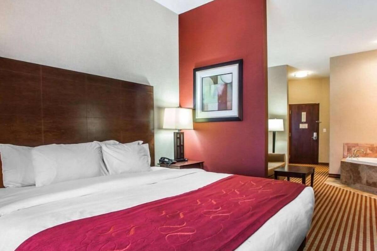 A hotel room with a large bed, a red accent wall, a framed picture, a desk with a lamp, and a bathroom visible in the background.