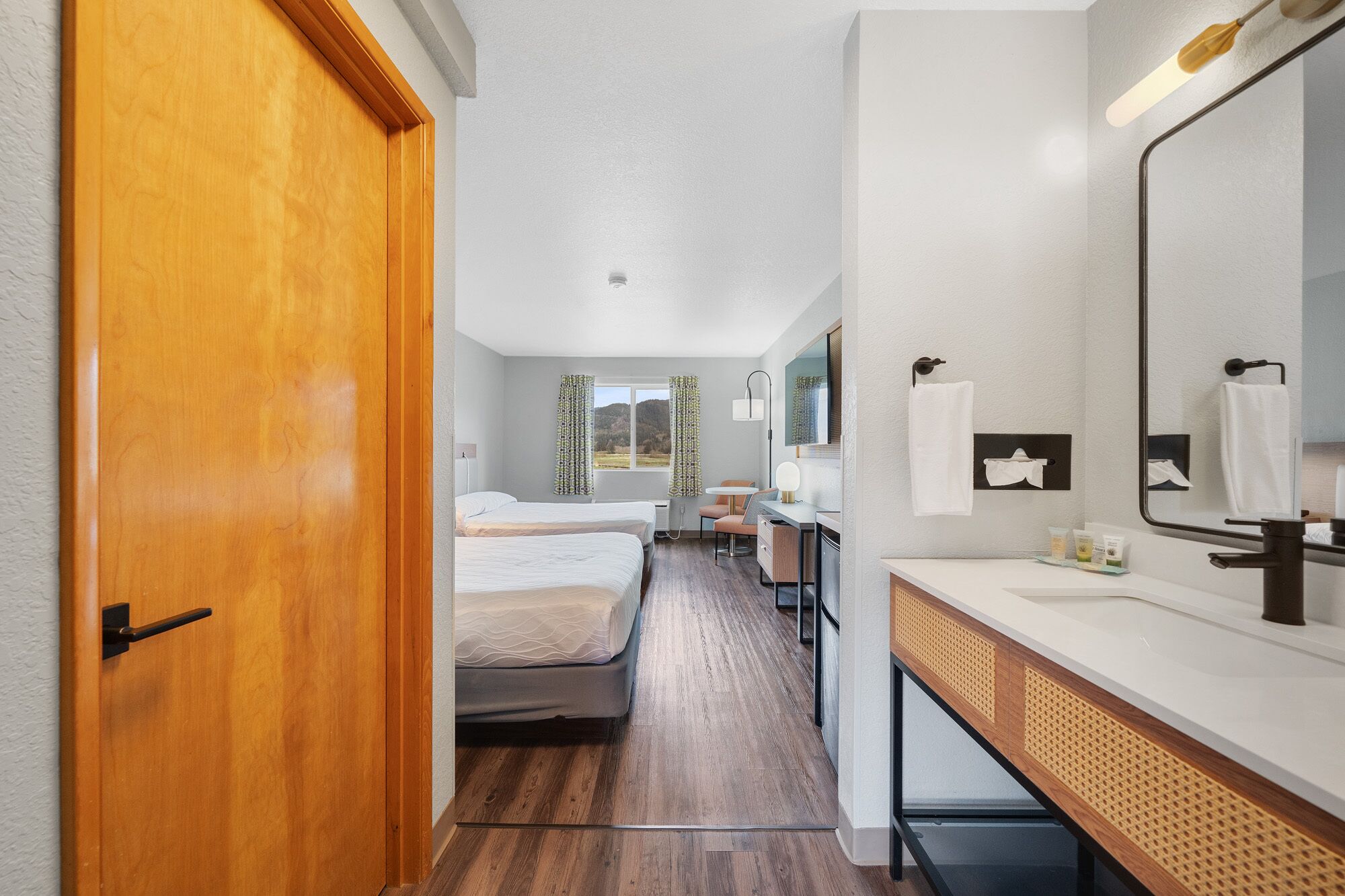 A hotel room with a wooden door, a bed, a desk with a mirror, a small table, and a window with curtains.