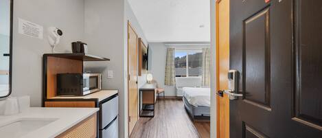 A hotel room with a kitchenette, a bed, a desk, and a bathroom.