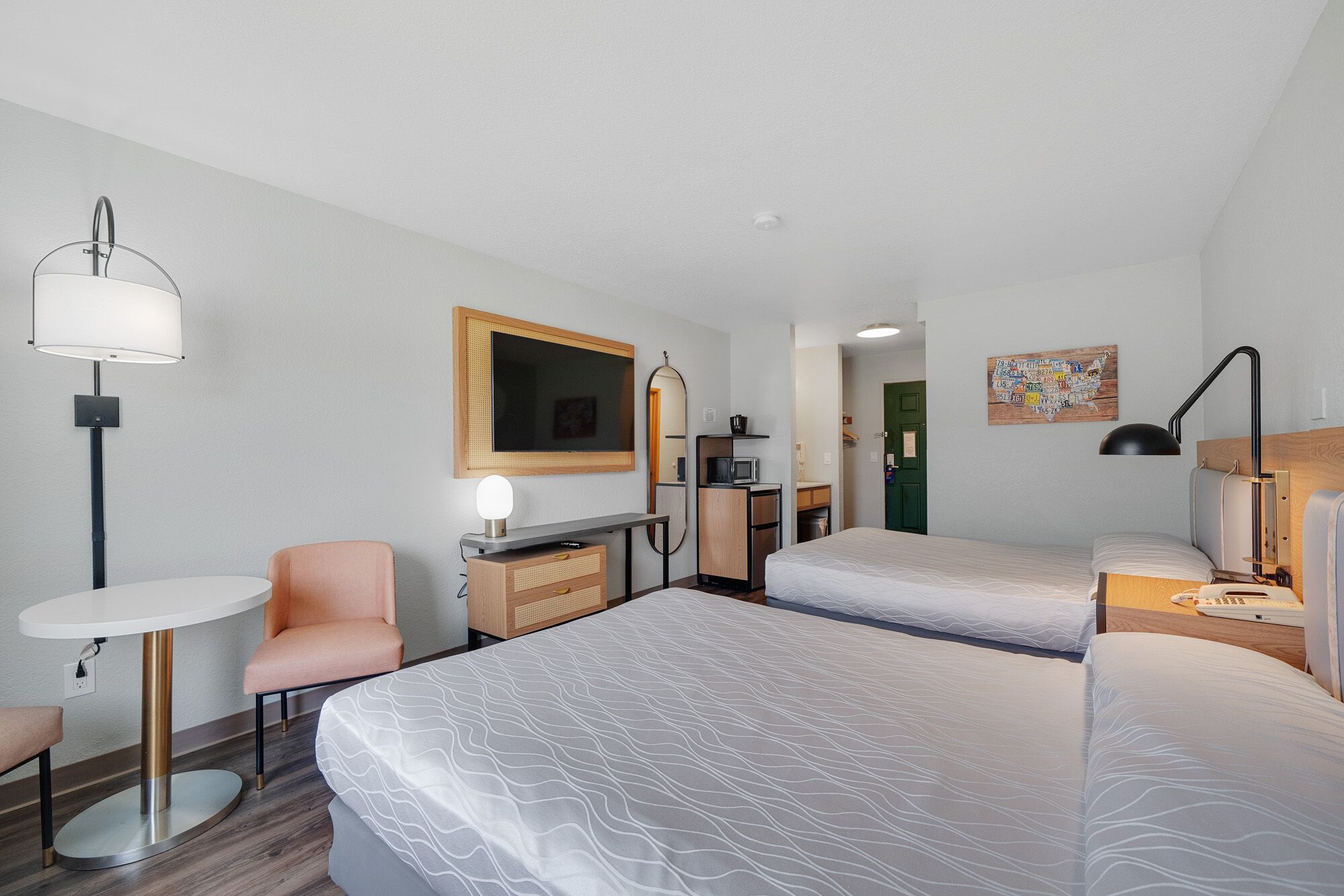 A hotel room with two beds, a desk, a chair, a lamp, a small table, and a wall-mounted TV.