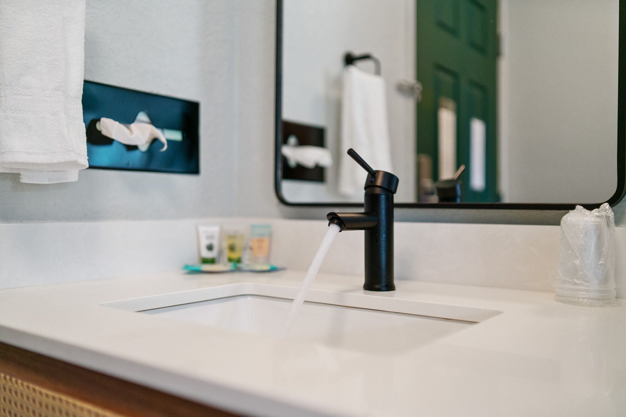 A modern bathroom with a black faucet, a white sink, and a mirror reflecting a towel and a green door.