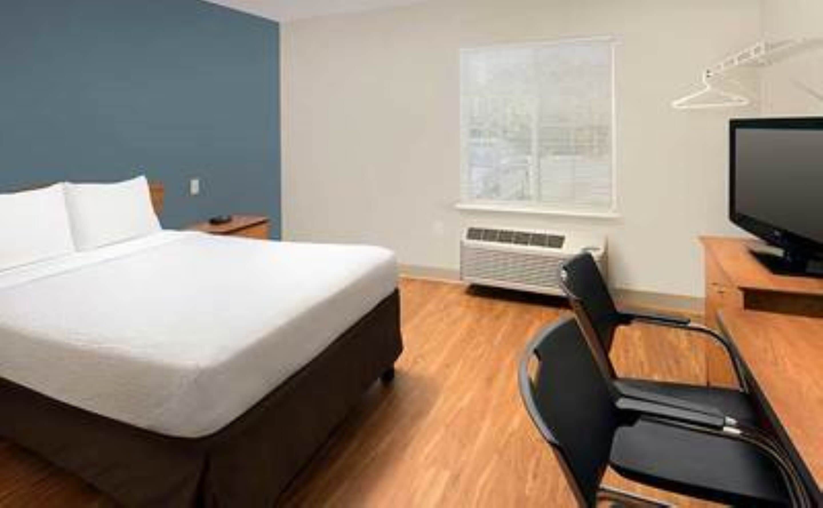 Habitación de hotel con cama, escritorio, silla, televisor y ventana con persianas.