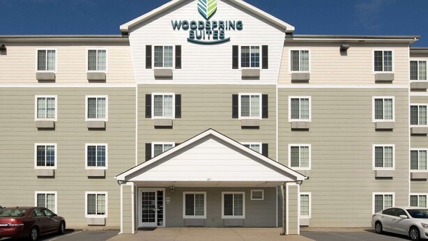 Un hôtel à plusieurs étages, doté d’une entrée principale et d’une enseigne indiquant « Woodspring Suites ».