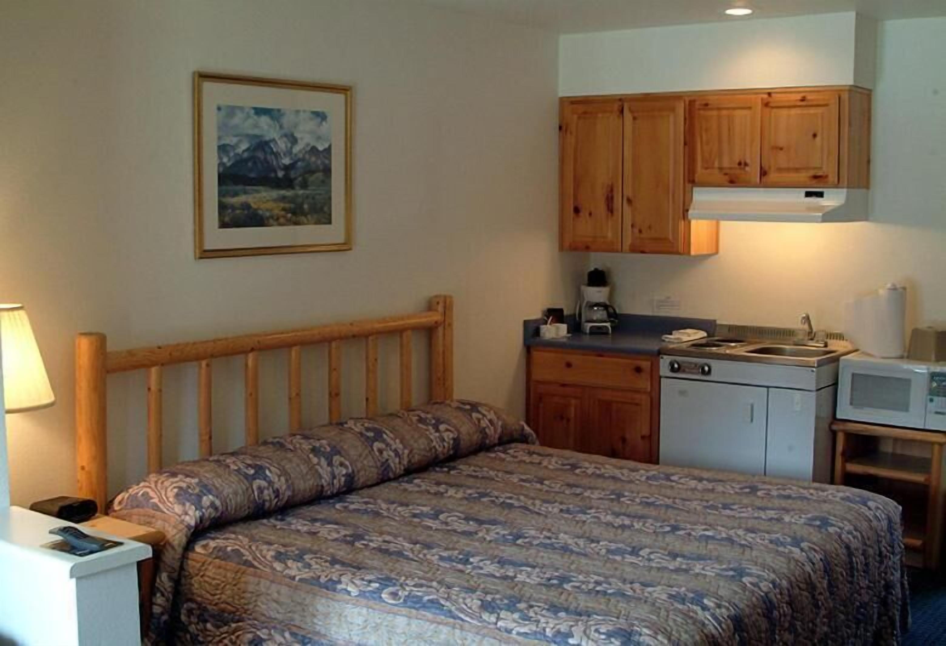 A bedroom with a bed, wooden headboard, a small kitchenette with a stove, microwave, and cabinets.