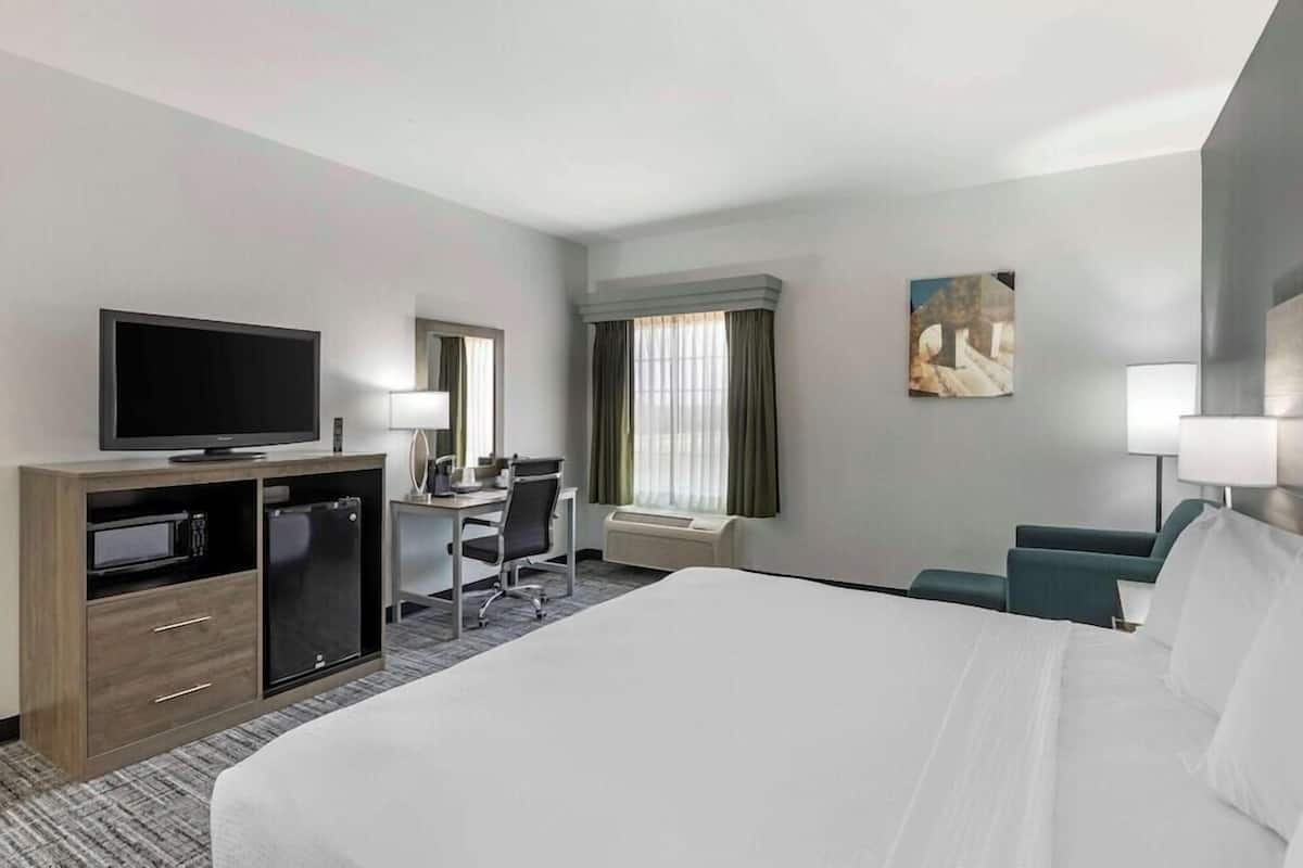 A hotel room with a large bed, a desk with a chair, a television, a mini-fridge, and a window with curtains.
