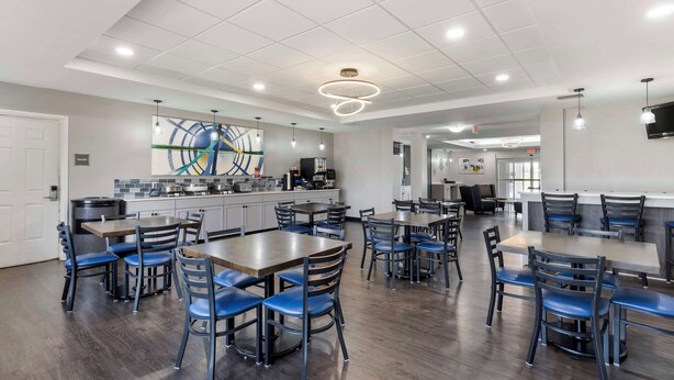 Un espace repas moderne doté de plusieurs tables et chaises, un coin cuisine équipé d’appareils électroménagers, et un plafond avec des spots encastrés et un luminaire circulaire.