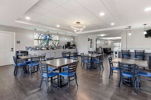 A modern dining area with multiple tables and chairs, a kitchen area with appliances, and a ceiling with recessed lighting and a circular light fixture. - Best Western Plus MidAmerica Hotel (Mascoutah)
