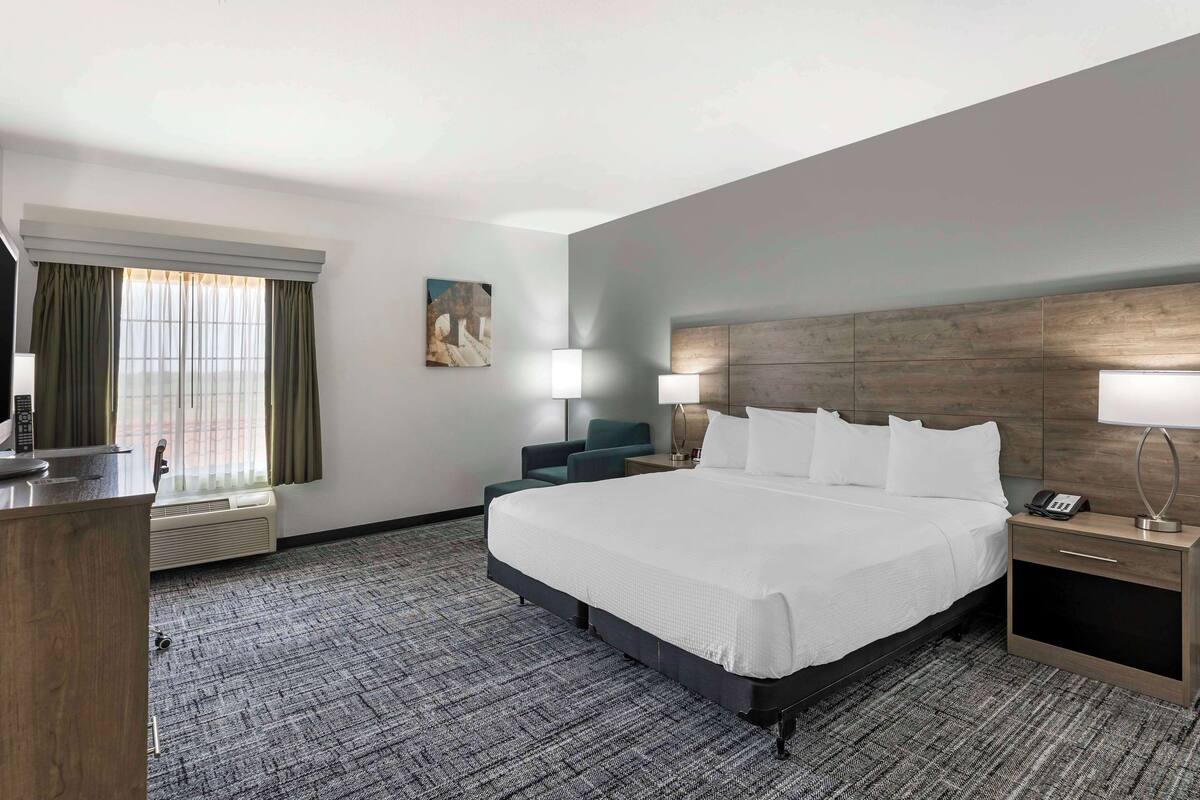 A hotel room with a large bed, a desk, a chair, a bedside table with a lamp, and a window with curtains.