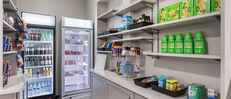 A hotel kitchenette with a refrigerator, shelves stocked with beverages and snacks, and a coffee machine.