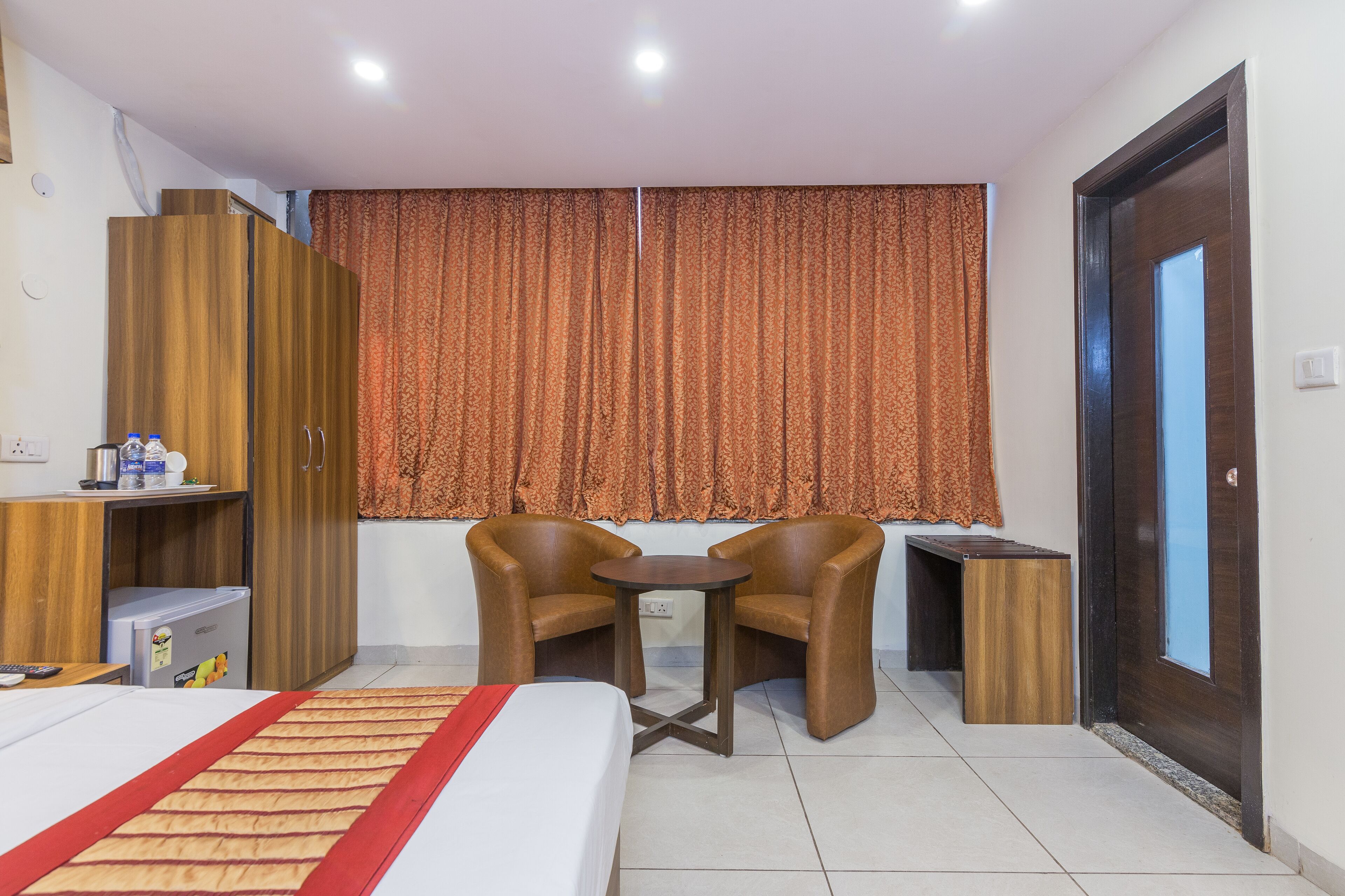 Quarto de hotel com cama, duas cadeiras, uma mesinha, guarda-roupa e janela com cortinas.