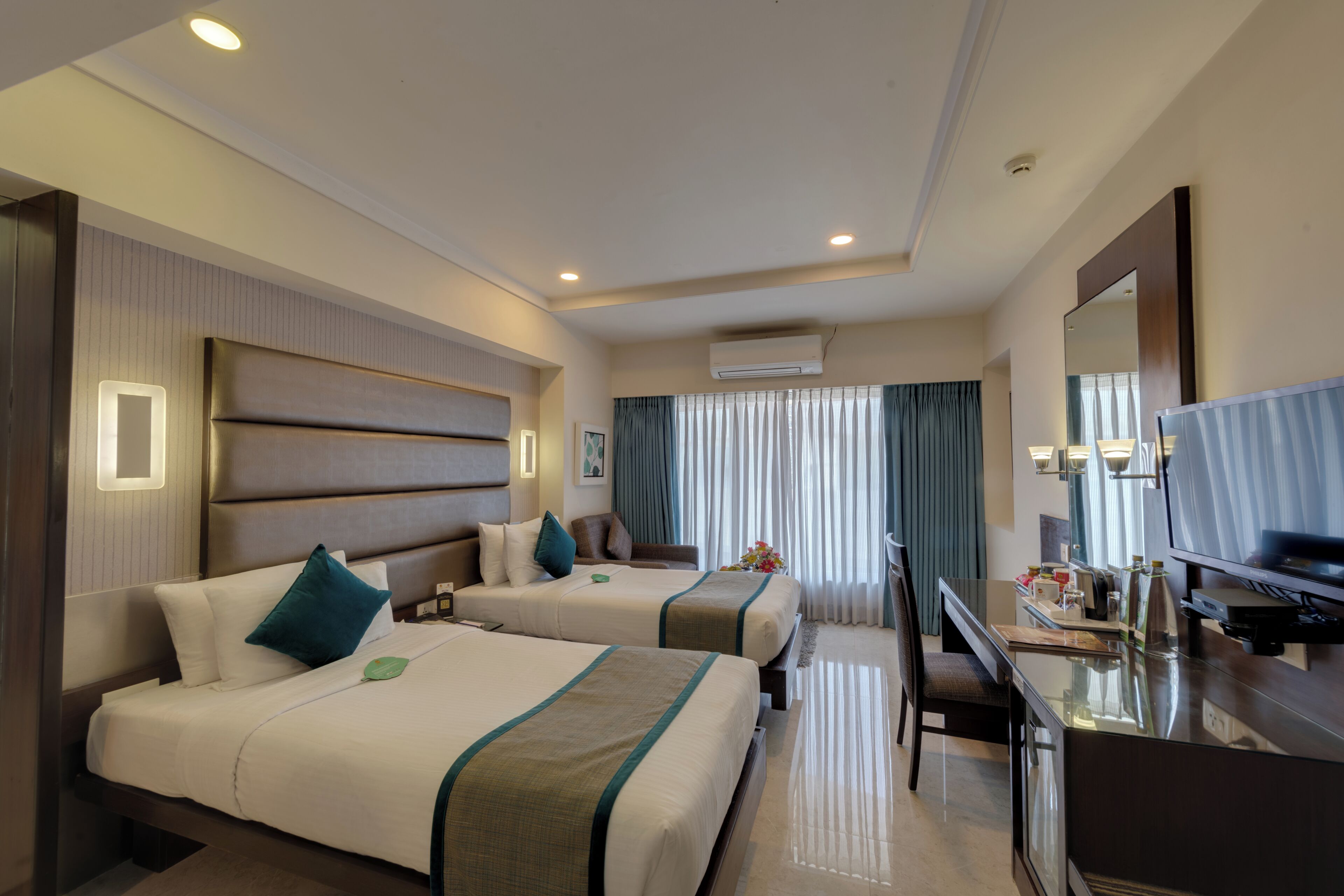 A hotel room with two beds, a desk, a TV, and an air conditioning unit.