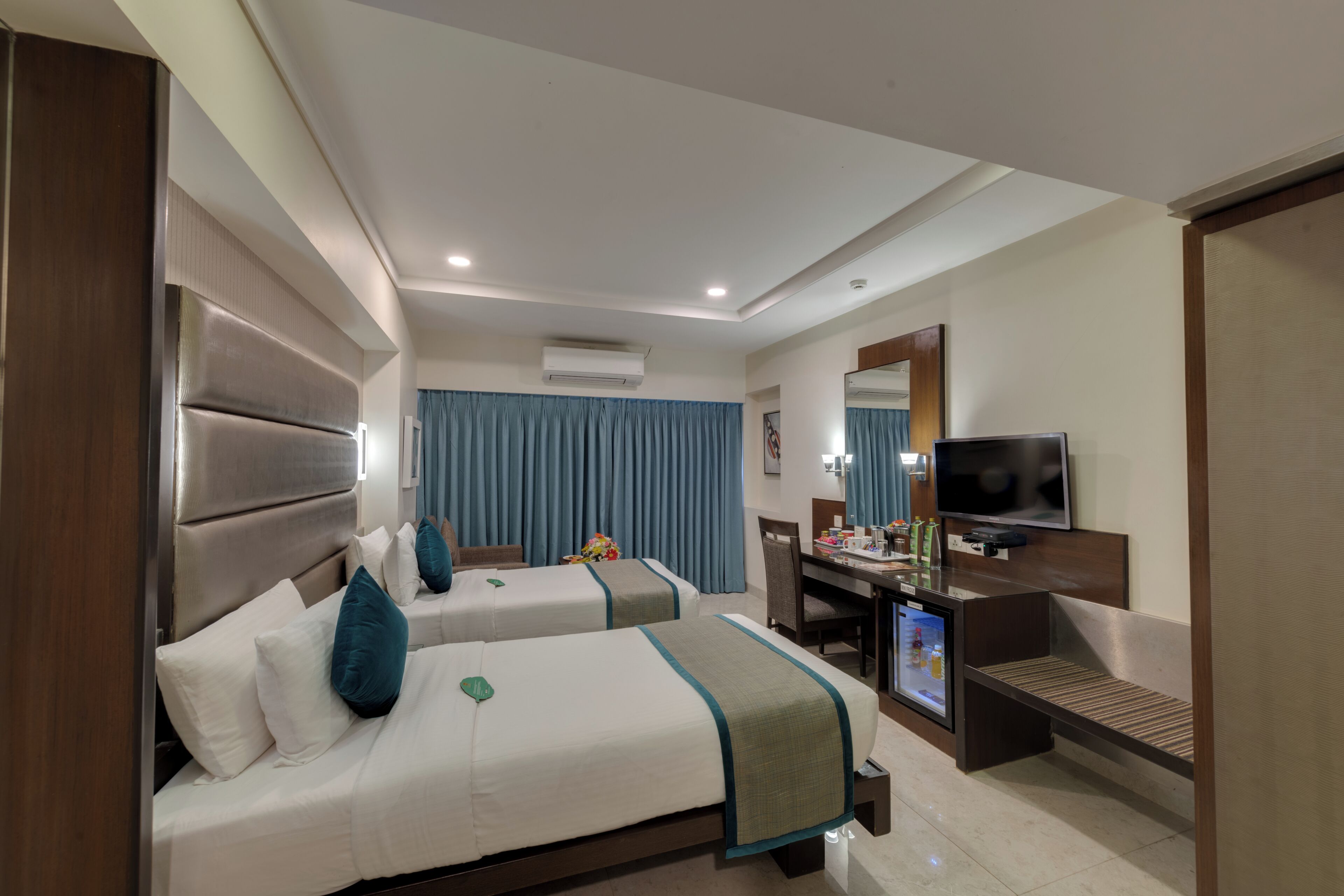 A hotel room with two beds, a desk with a TV, and an air conditioning unit.