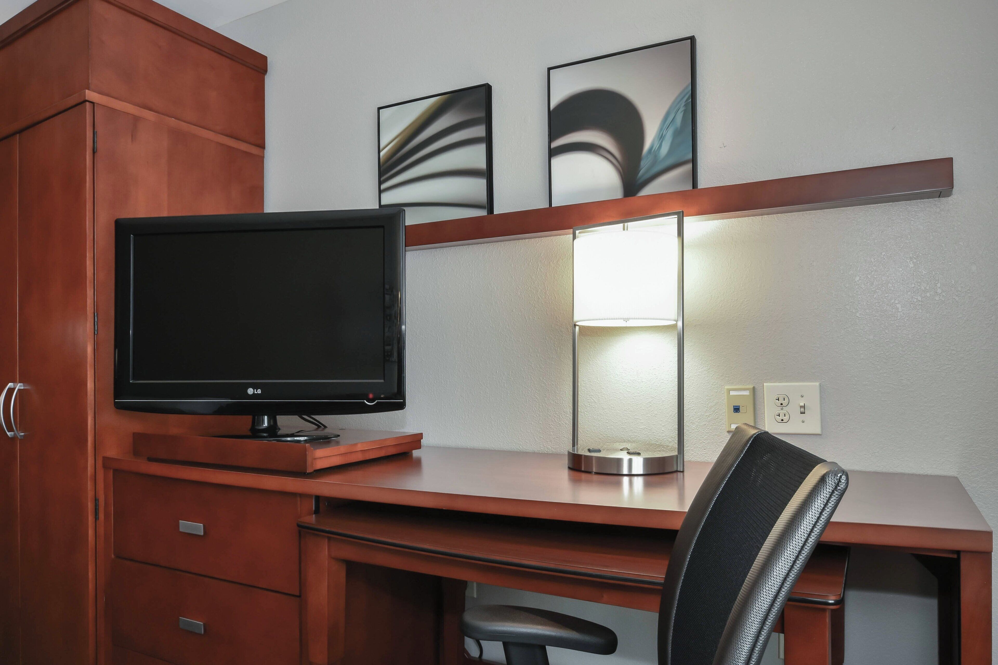 A hotel room with a wooden desk, a flat-screen TV, a lamp, and a chair.