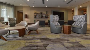 A modern hotel lobby with a carpeted floor, seating area with sofas and armchairs, a reception desk, and a wall-mounted shelf with decorative items. - Courtyard Marriott Concord (Concord)