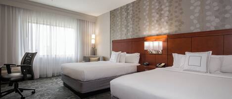 Quarto de hotel com duas camas, uma escrivaninha, uma cadeira, um criado-mudo e um papel de parede estampado.