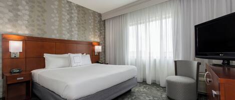 A hotel room with a large bed, a television, a chair, and a desk with a lamp.