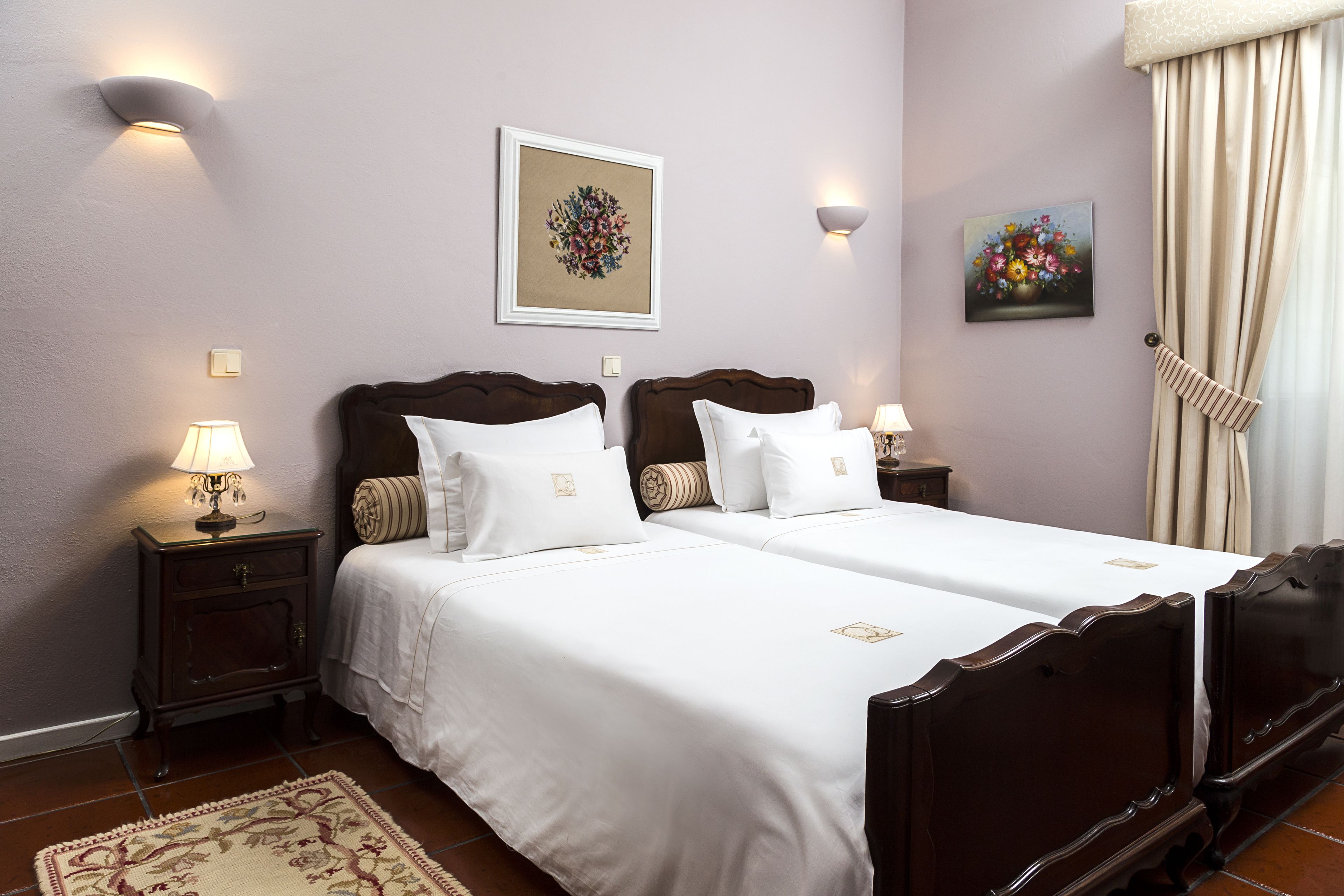 A hotel room with two beds, a nightstand with a lamp, a framed floral artwork, and a painting of flowers.