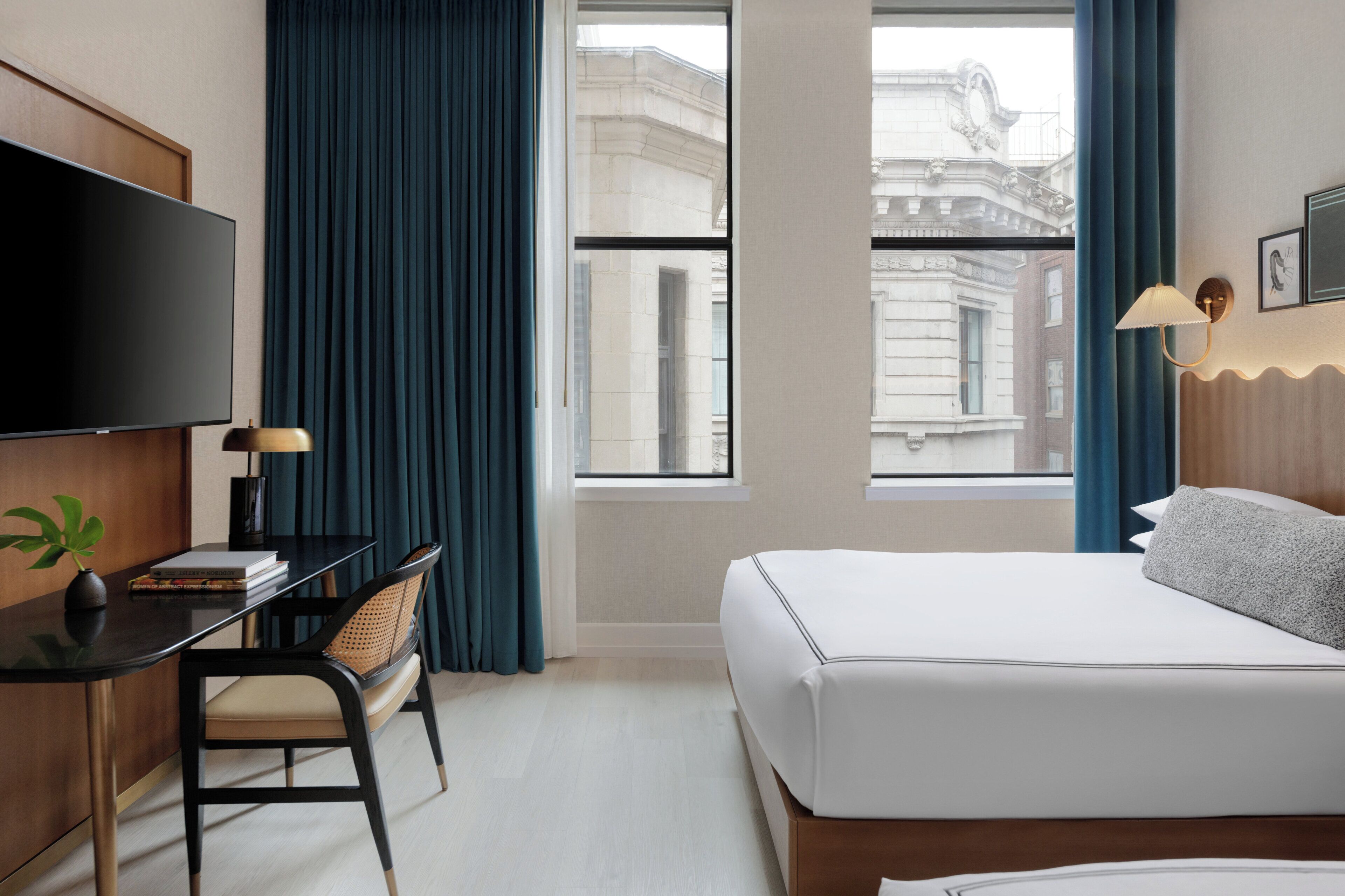 A hotel room with a bed, a desk with a chair, a television, and a window with curtains.