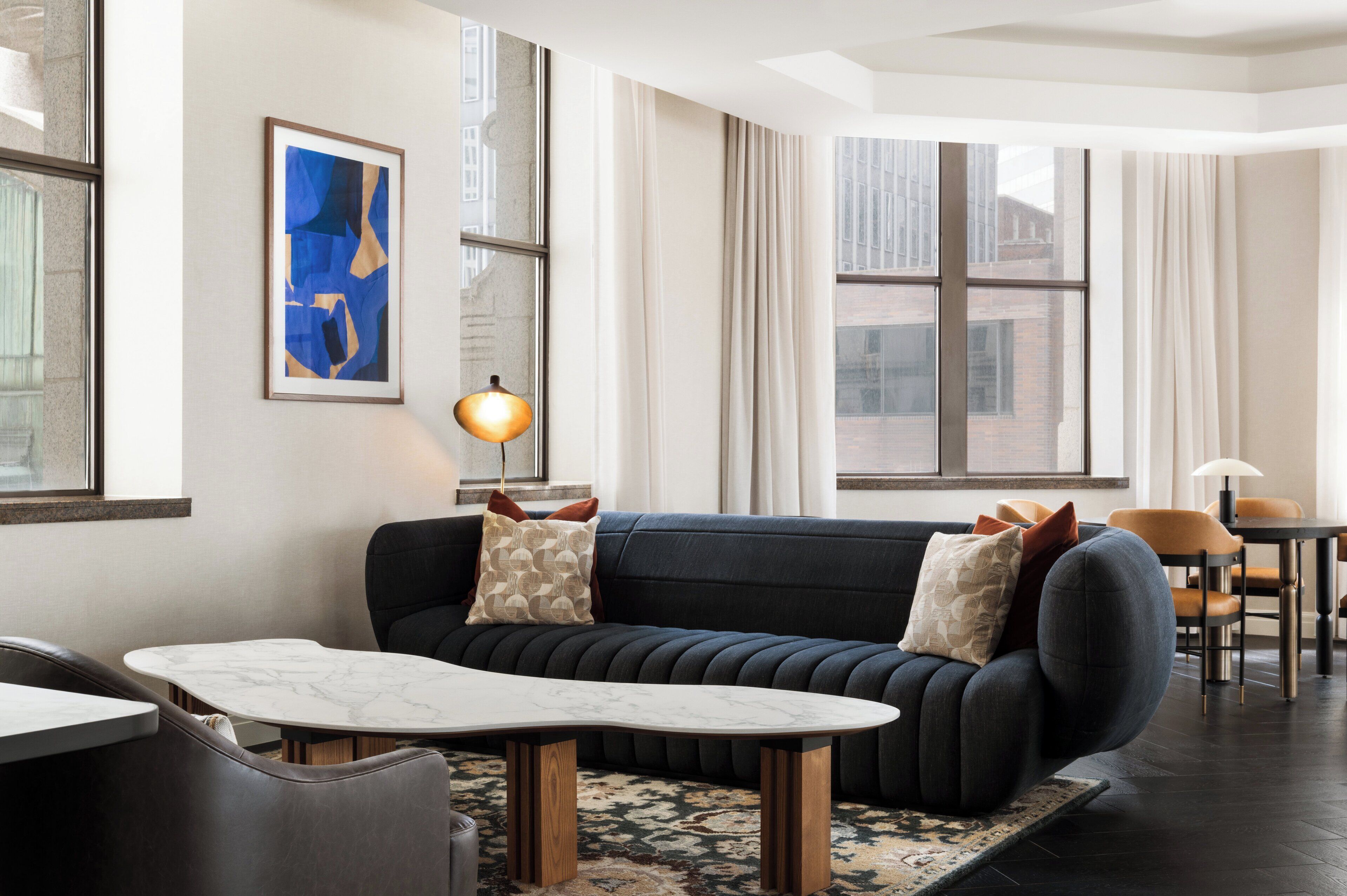 A modern living room with a dark blue sofa, a marble coffee table, and a patterned rug. There is a dining area with a small table and chairs, and a large window with curtains.