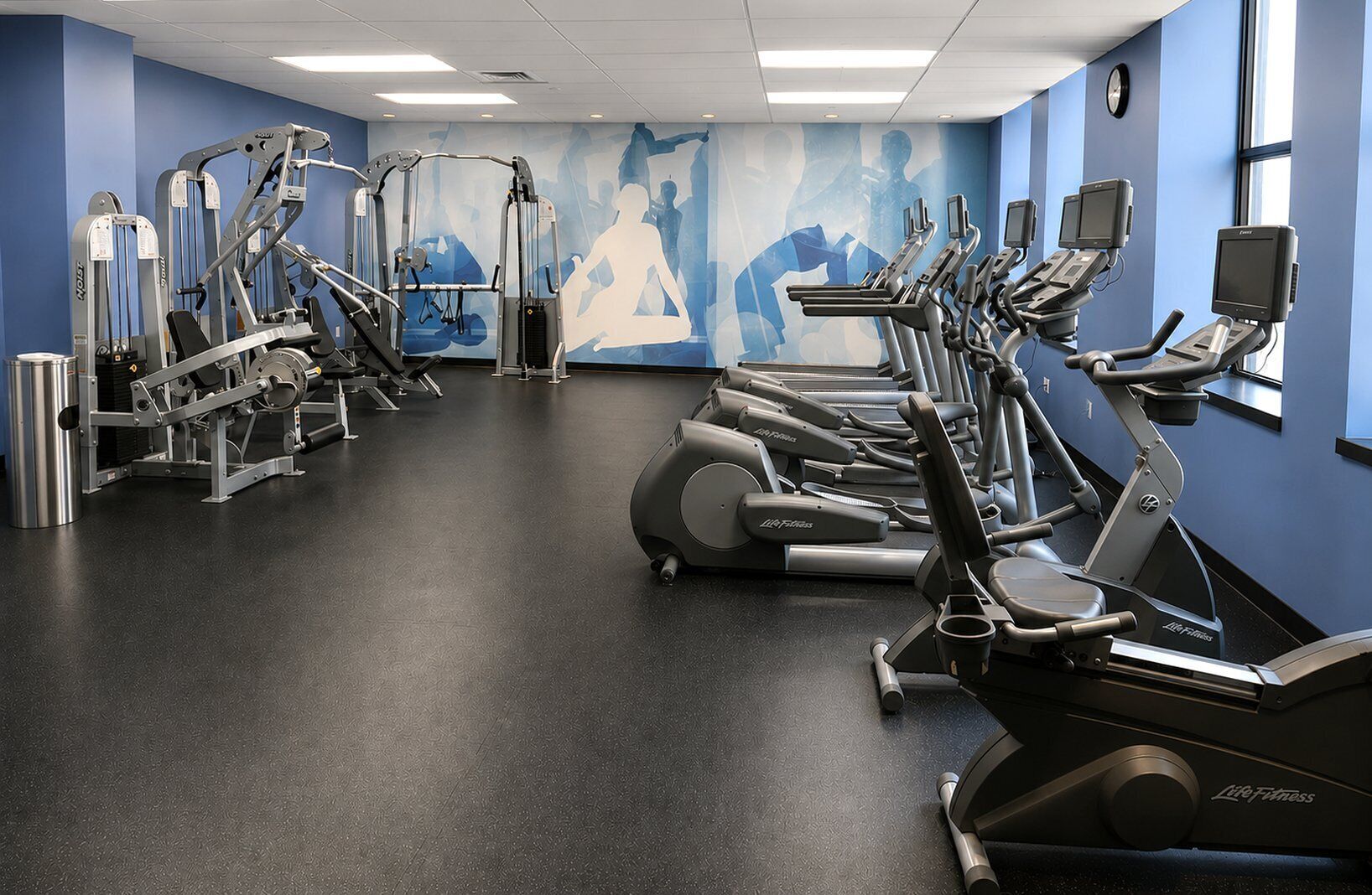 A gym with cardio machines, weight machines, and a mural on the wall.