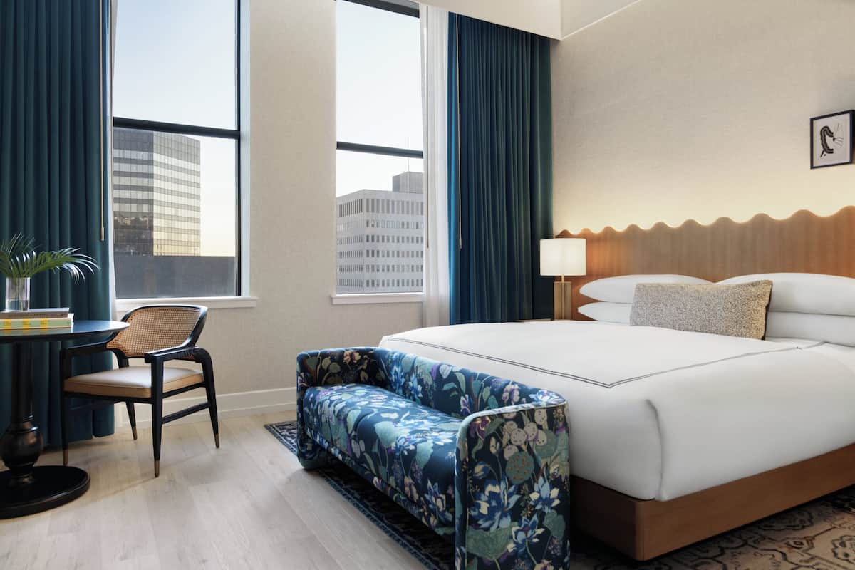 Habitación de hotel con una cama grande, zona de estar, escritorio con silla y vistas a la ciudad a través de las ventanas.