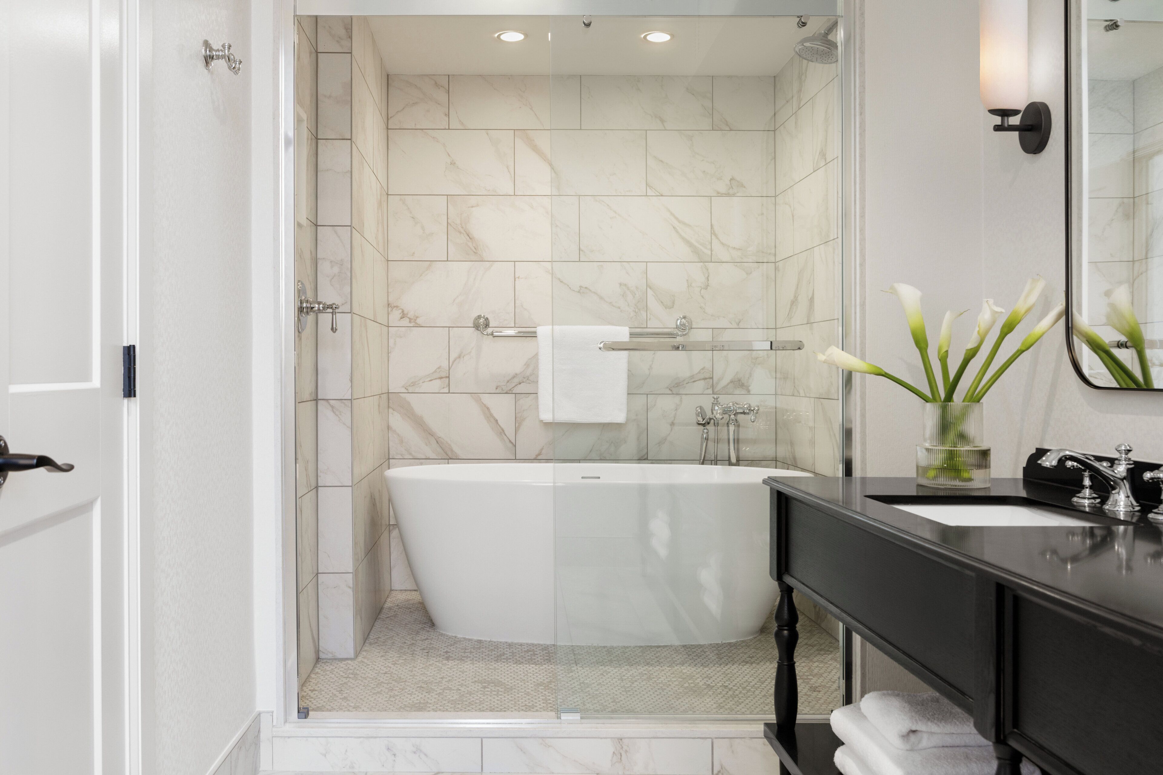 A modern bathroom with a freestanding bathtub, a glass shower enclosure, a vanity with a mirror, and a vase with flowers.