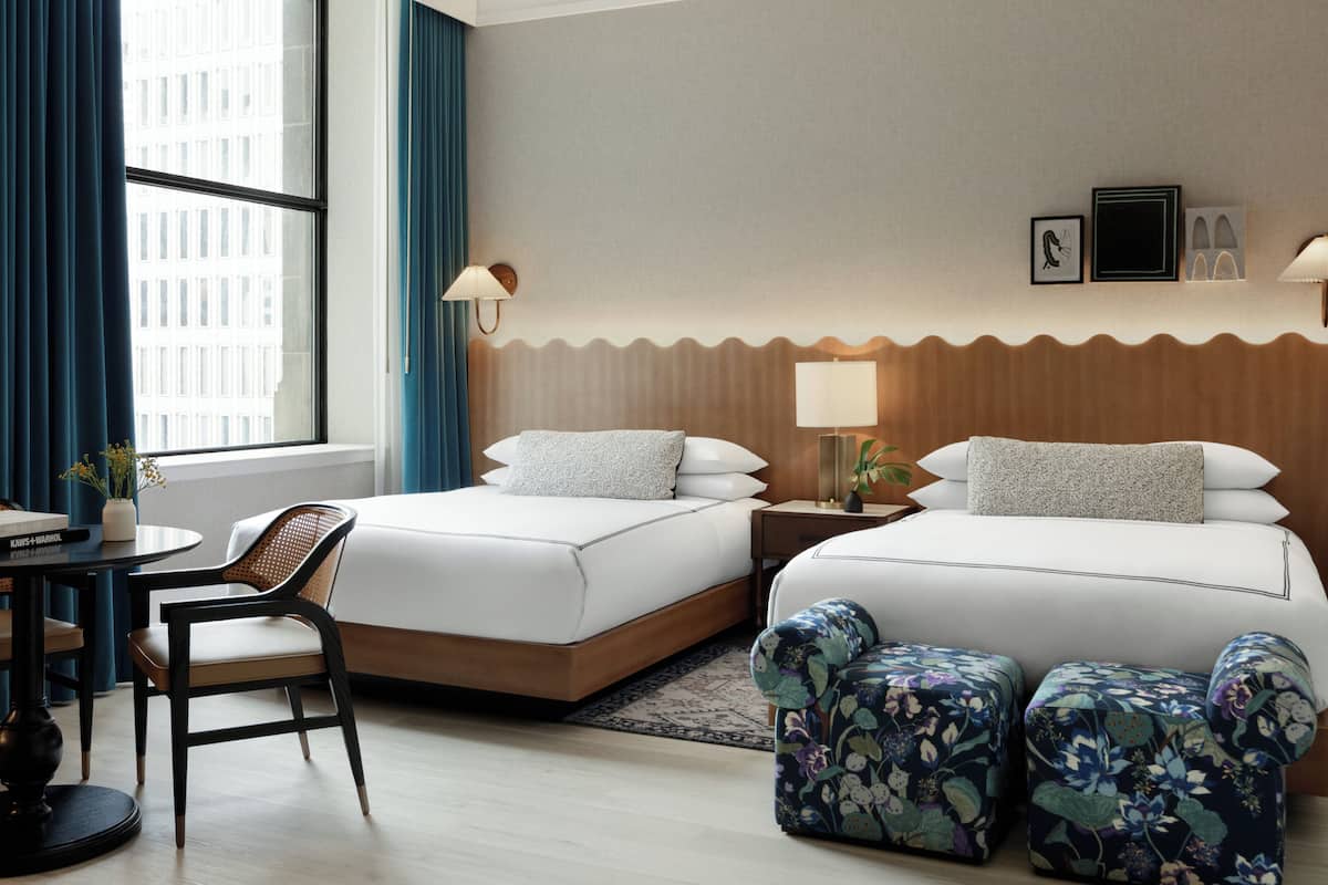 Una habitación de hotel con dos camas, un escritorio, una silla y un ventanal con cortinas.