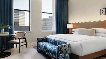 A hotel room with a large bed, a seating area, a desk with a chair, and a view of the cityscape through the windows.