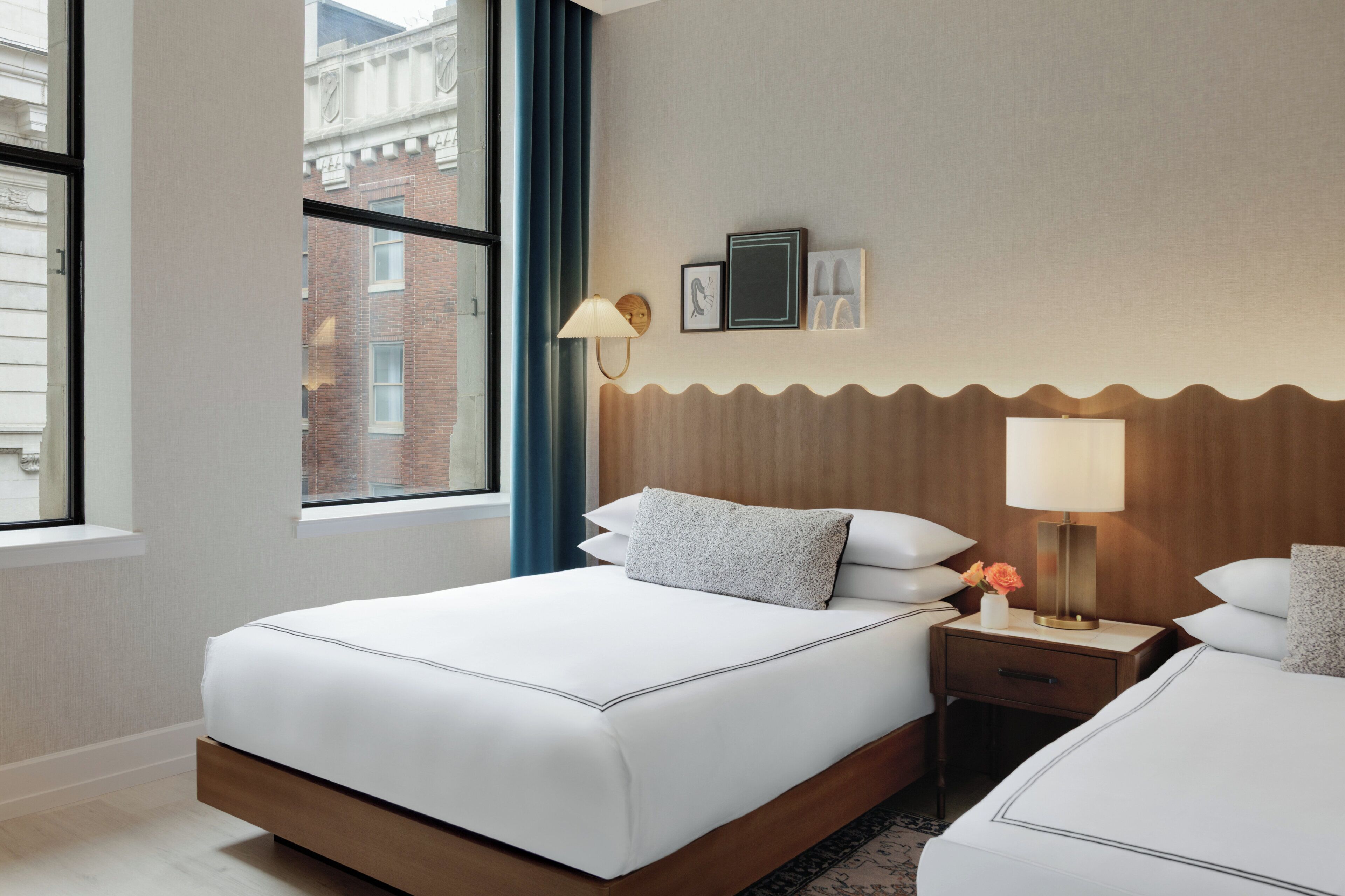 A hotel room with a large bed, a nightstand with a lamp, a wall-mounted picture, and a window with a view of a building.