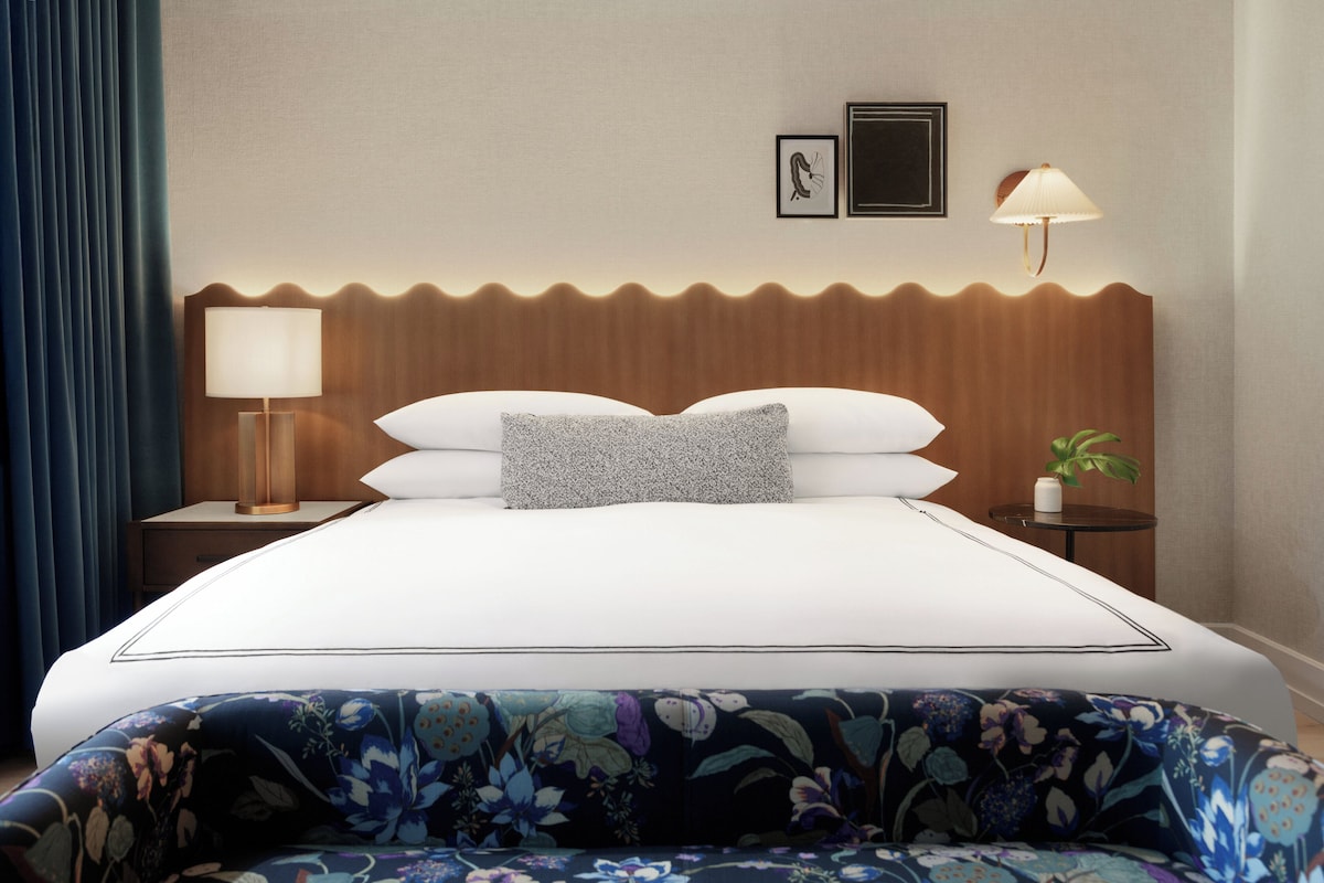 A hotel room with a bed, a nightstand, a lamp, a wall-mounted picture, and a small table with a plant.