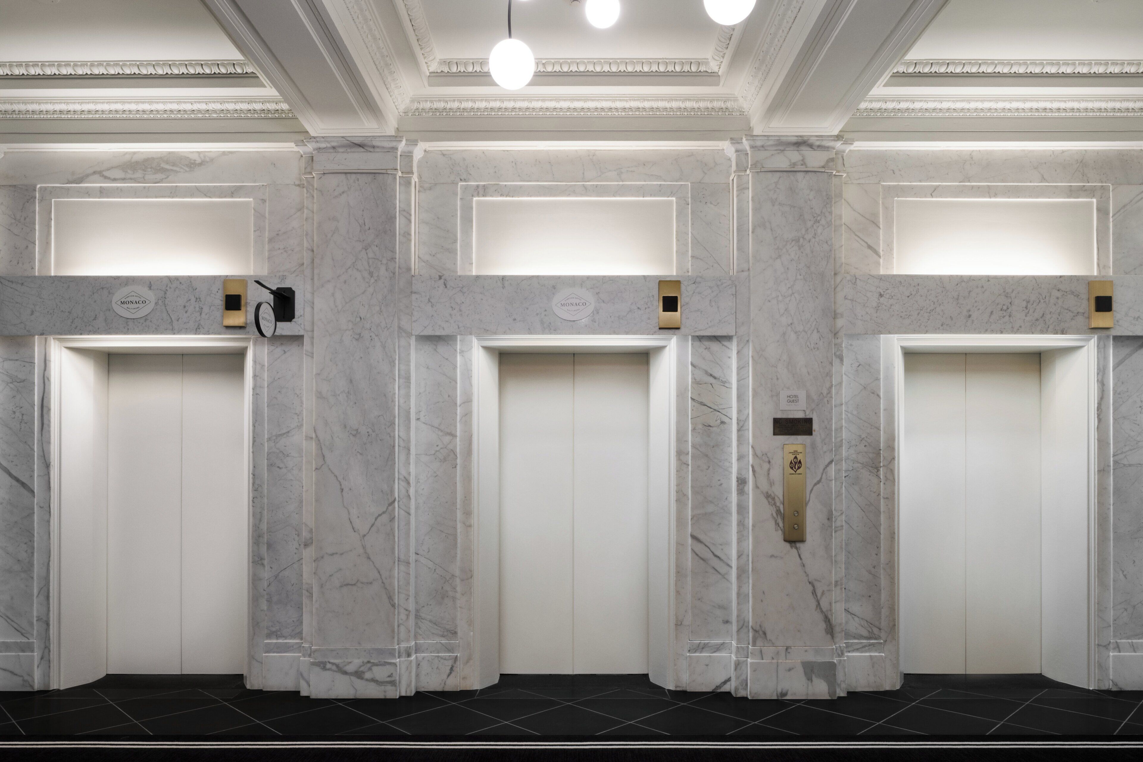 Un vestíbulo de hotel con paredes de mármol, tres ascensores y un suelo de baldosas negras.