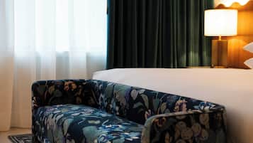 A hotel room with a bed, a nightstand with a lamp, a floral-patterned armchair, and sheer curtains.