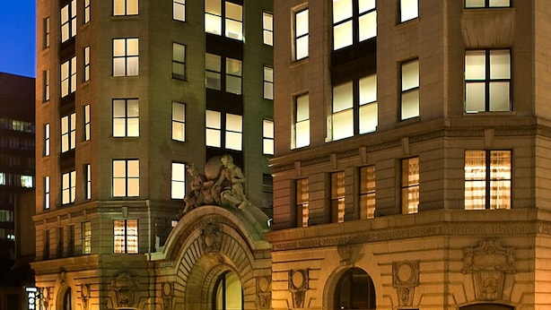 Ein mehrstöckiges Hotel mit beleuchteten Fenstern, einem markanten Eingang mit Vordach und einer Statue darüber.