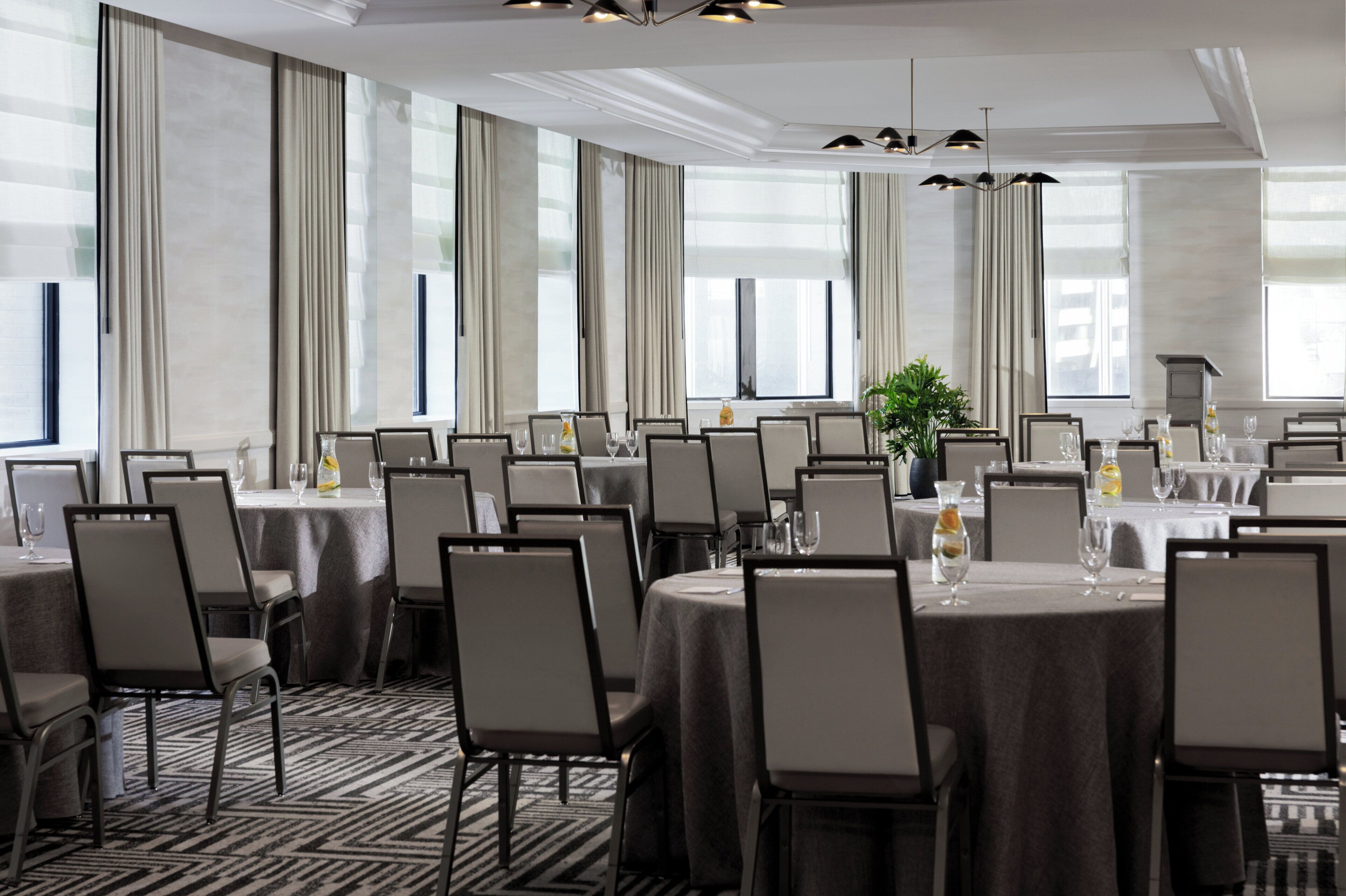 A conference room with multiple tables and chairs, large windows with curtains, and a modern chandelier.
