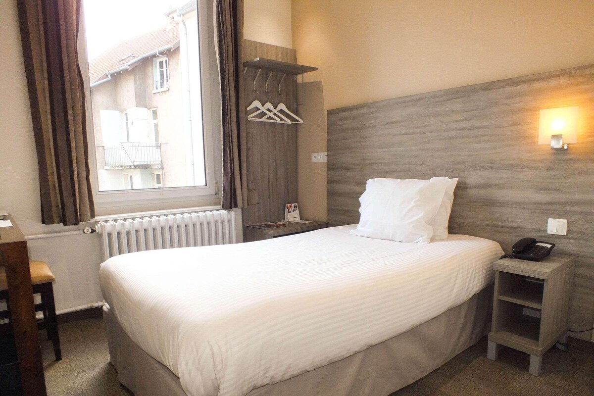 A hotel room with a bed, bedside table, lamp, and a view of the outside.
