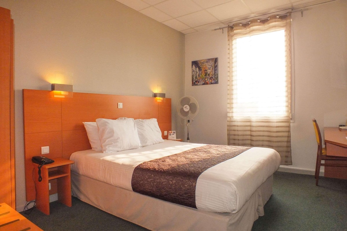 A hotel room with a bed, bedside table, chair, and a fan.