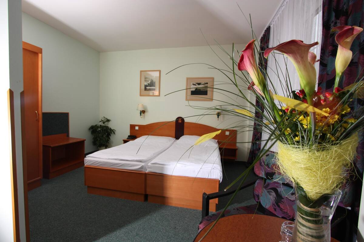 A hotel room with a bed, a desk, a chair, a lamp, and a vase with flowers.