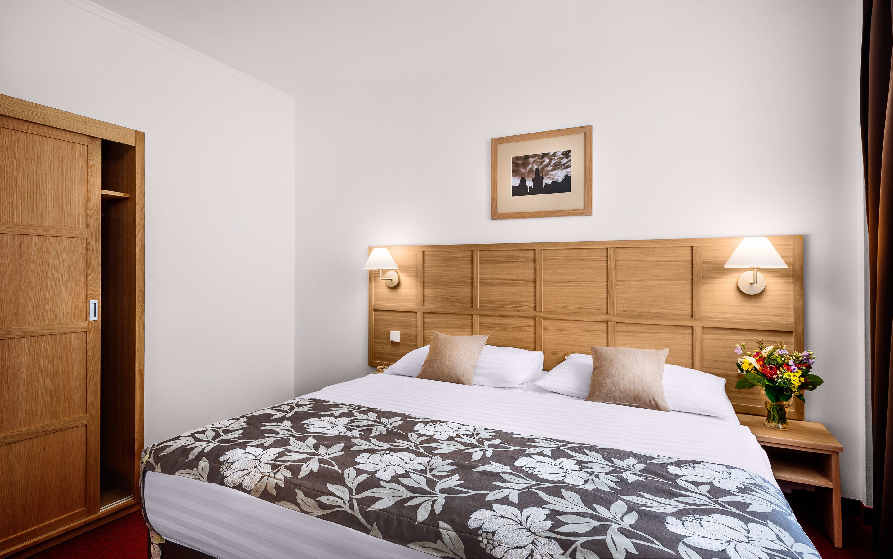 A hotel room with a wooden headboard, two bedside lamps, a bed with floral bedding, a nightstand with a vase of flowers, and a framed picture on the wall.