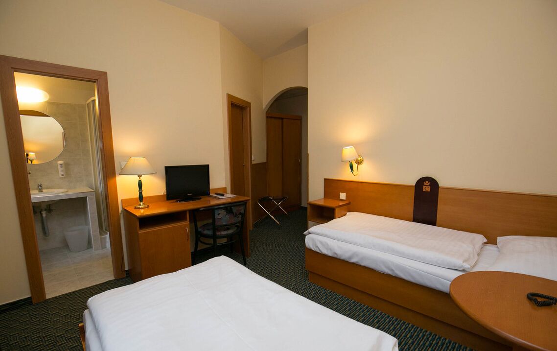Hotellihuone, jossa on kaksi sänkyä, työpöytä, jossa on tietokone, tuoli, pieni pöytä ja kylpyhuone, joka näkyy avoimesta ovesta.