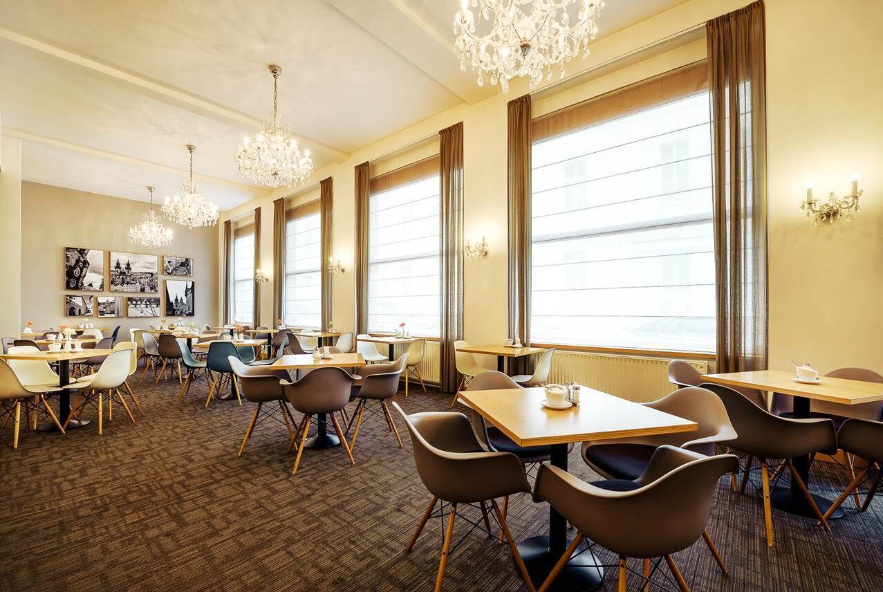 A hotel dining area with multiple tables and chairs, large windows with curtains, and chandeliers.