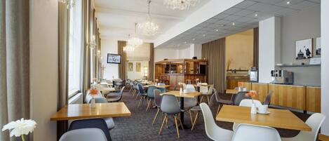 A hotel dining area with multiple tables and chairs, chandeliers, and a kitchen in the background.