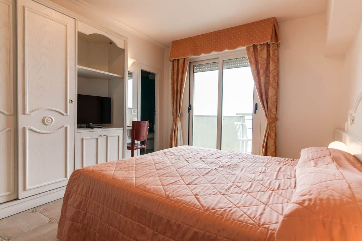 Une chambre avec un grand lit, une télévision, un fauteuil et un balcon avec des rideaux.