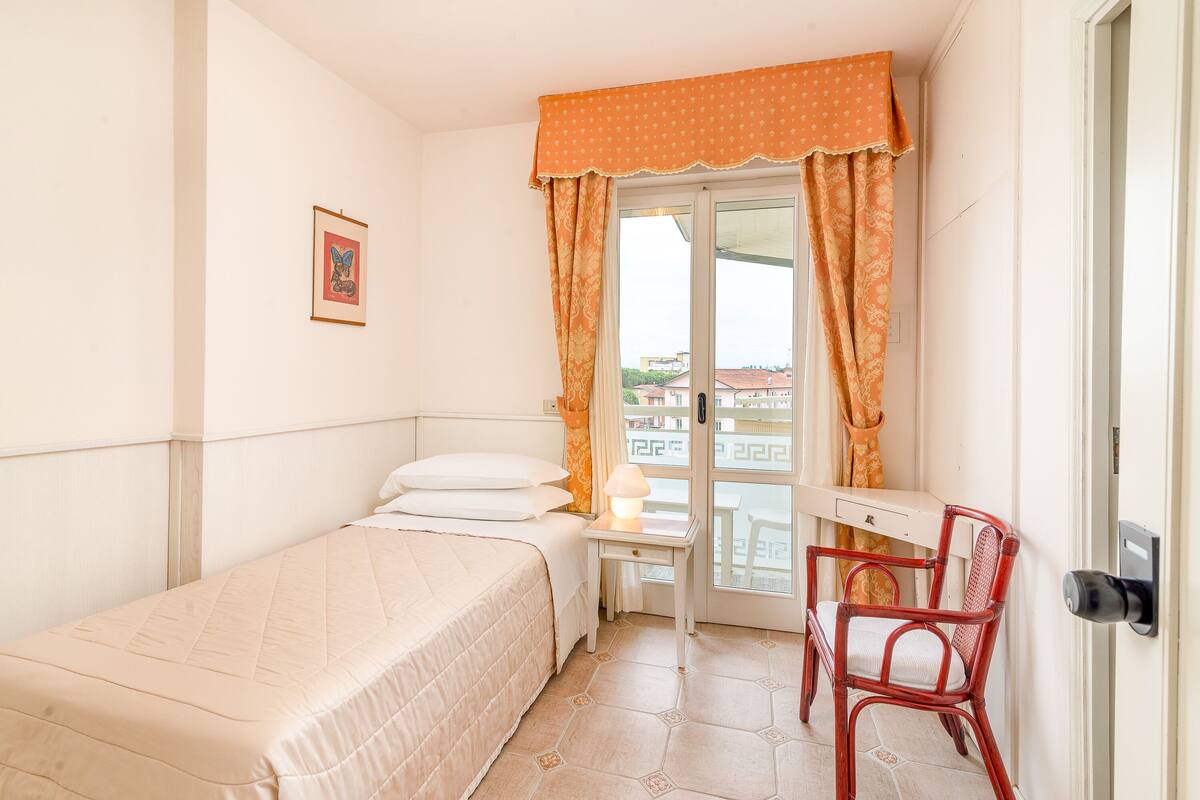 Un lit simple avec une couette, une table de chevet avec une lampe, une chaise et une fenêtre avec des rideaux.